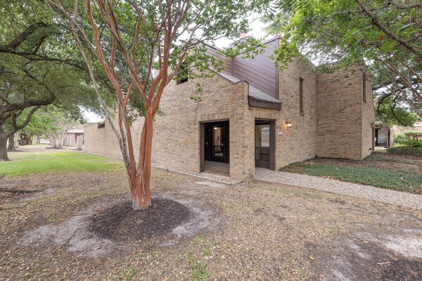 A brick building with a door and windows, surrounded by trees and a gravel path. - The Aggieland House _ Game Day Retreat _ Walk to Kyle Field_Convenient Parking (College Station)