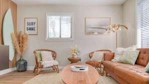 A living room with a brown leather sofa, two wicker chairs, a round wooden coffee table, a large window with blinds, and a surfboard decoration. - Luxury Villa 3 Blocks to Beach, Saltwater Pool, Outdoor Oasis, firepit, sunlounge's, mini putt and close to all the area has to offer. (Cape Canaveral)