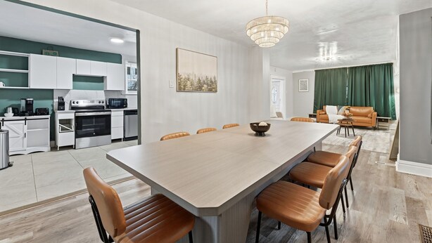 Una cocina moderna con armarios blancos, un comedor con una mesa larga y sillas marrones, y una sala de estar con un sofá verde y cortinas.