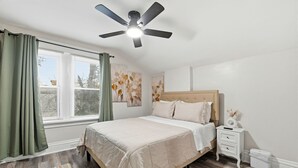 Un dormitorio con cama, mesita de noche, ventilador de techo, ventana con cortinas y un cuadro floral en la pared.