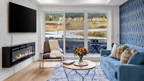 A modern living room with a fireplace, a sofa, a coffee table, and a large window with a view of a road and trees. - Brand New 1BR Canmore Retreat: Hot Tub, Sauna, Gym (Canmore)