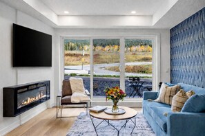 Ein modernes Wohnzimmer mit Kamin, einer Couch, einem Couchtisch und einem großen Fenster mit Blick auf eine Straße und Bäume.