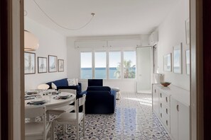 Ein Zimmer mit Esstisch für vier Personen, einer Couch, einem Sessel und Meerblick durch große Fenster.