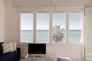 A room with a large window overlooking the sea, a television on a stand, and a dark blue sofa with patterned cushions. - CASA MEDITERRANEA - Vista Mare - Parcheggio (Sanremo)