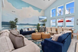 A living room with a beach mural, a dining area with a view, and a large window with a red umbrella. - Beachfront | Swim Spa | Game Room | Sleeps 15 (Galveston)