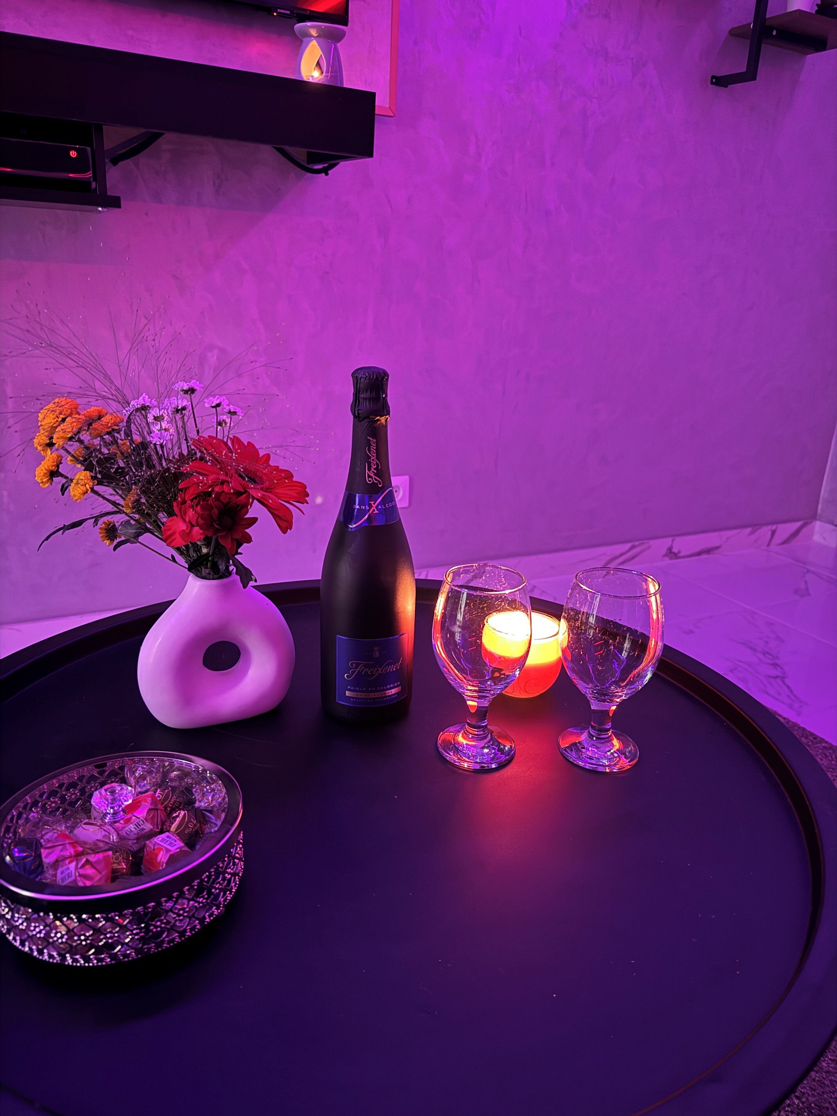 A table with a bottle of champagne, a vase of flowers, two glasses, and a candle.