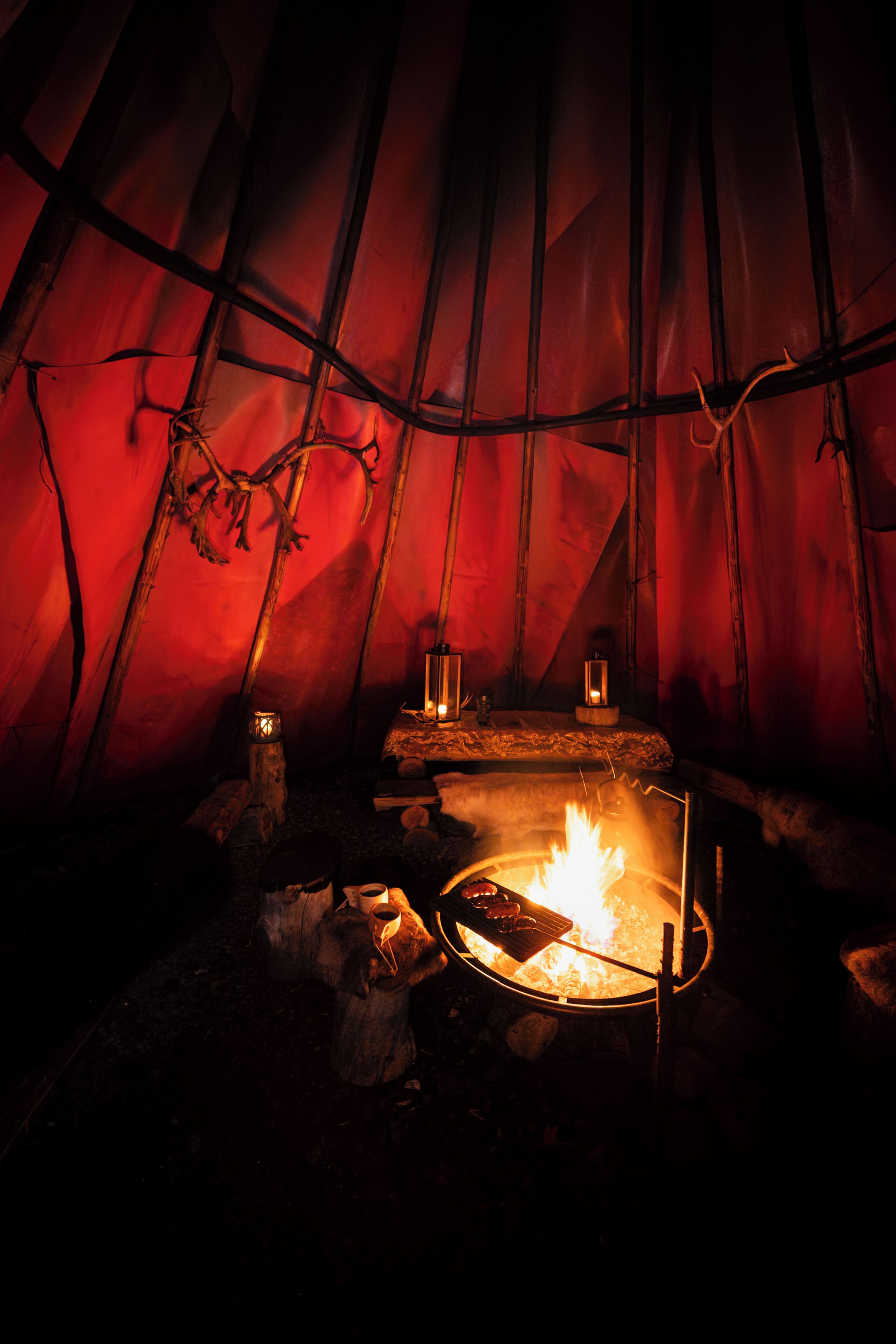 A tent interior with a central fire, wooden branches, and lanterns.