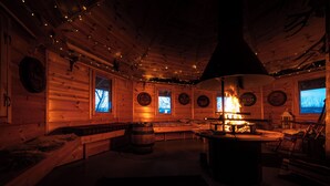 Interior acogedor de una cabaña con paredes de madera, una chimenea central y ventanas con vistas al exterior.