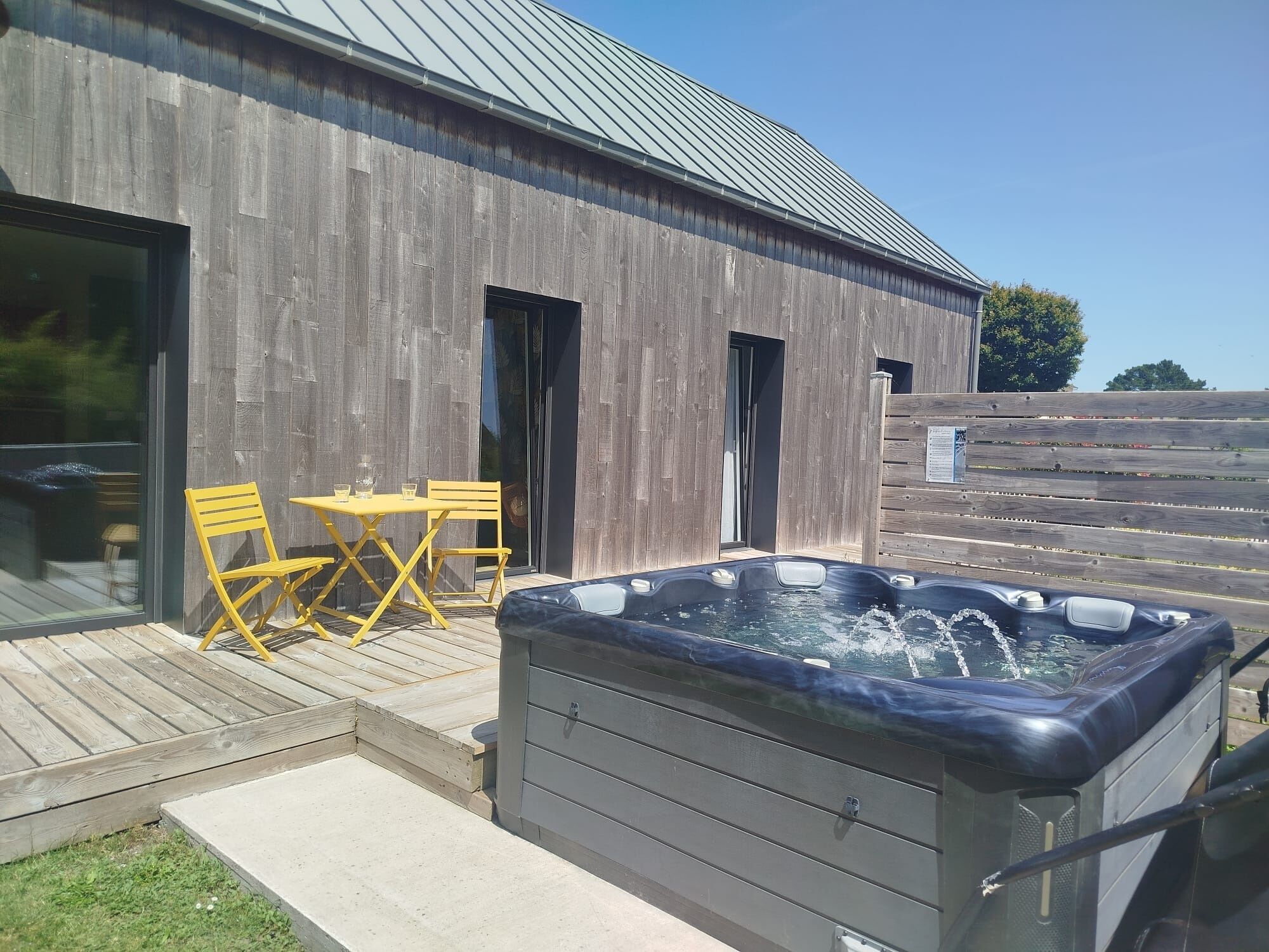 A wooden building with a hot tub, a table, and two chairs on a wooden deck.
