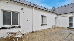 Ein weißes Gebäude mit einer mit Fliesen ausgelegten Terrasse, einem Metalltisch und -stühlen sowie einem Fenster mit Rollläden.