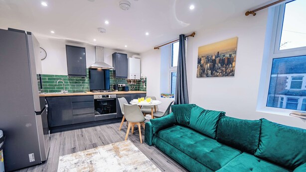 A modern kitchen with a dining area, a green sofa, and a cityscape view. - Modern 2BR Flat Steps from Cardiff Pubs & Town (Cardiff)