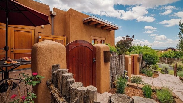 A rustic building with a wooden door, a red umbrella, and a dining table with food. - Luxury Adobe Retreat with Jemez Mountain Views (Santa Fe)