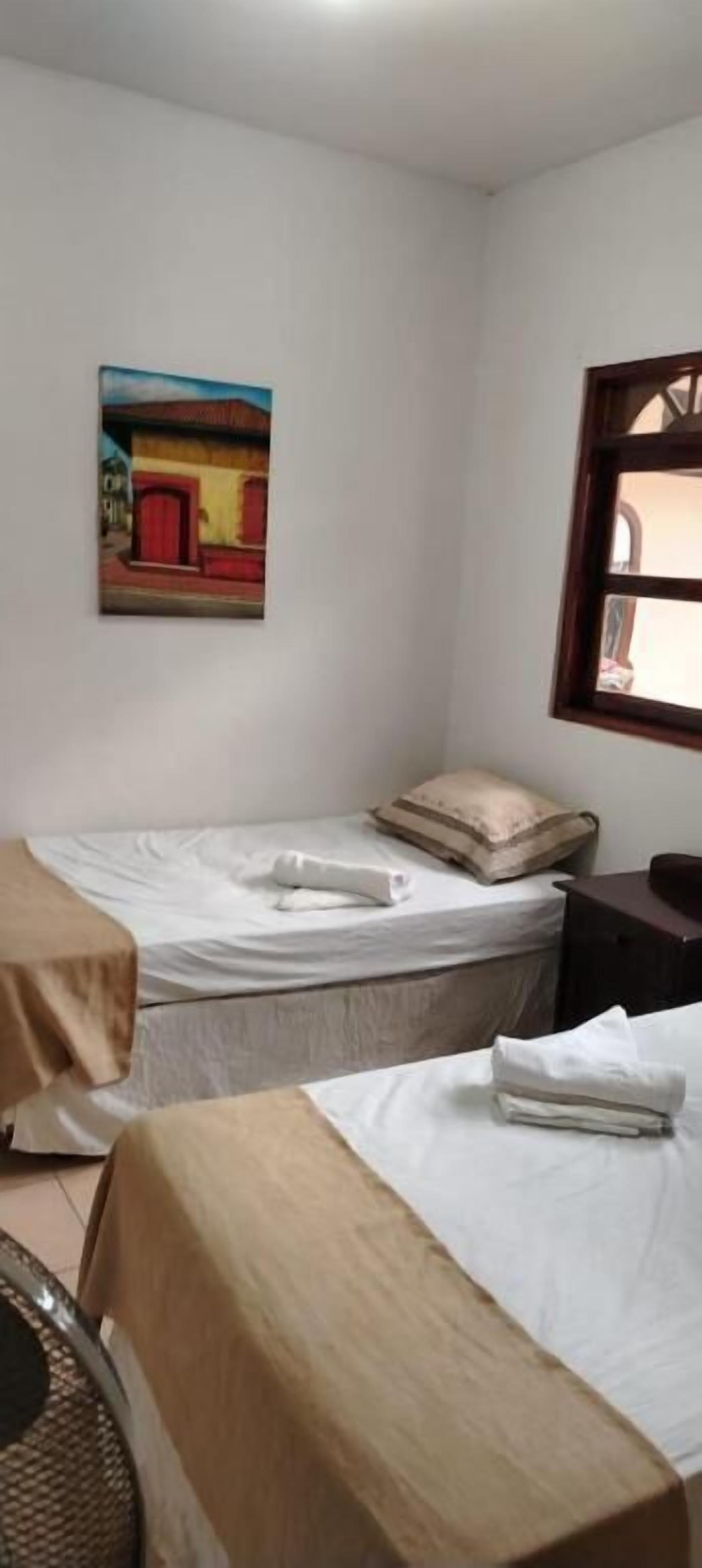 A small, simple room with two single beds, a wooden chair, a small table, a painting on the wall, and a window with a wooden frame.