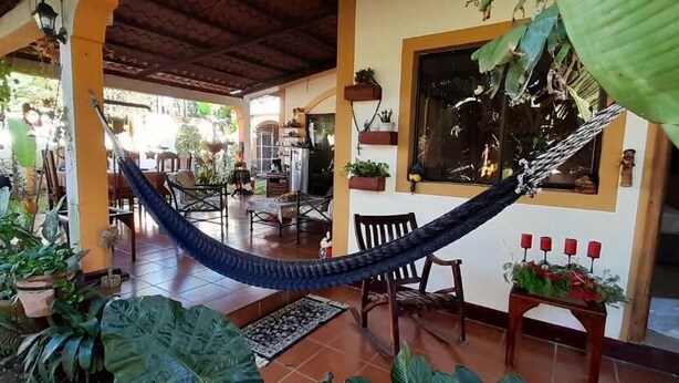 Een hangmat in een overdekte buitenruimte met stoelen en tafels, omgeven door potplanten en een betegelde vloer.