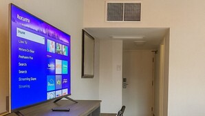A hotel room with a wooden desk, a television displaying a channel guide, a mirror, a bed, and a door leading to another room. - Two Queen Beds (Non Smoking) (Point Edward)
