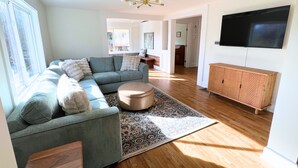 A modern living room with a sectional sofa, a round ottoman, a patterned rug, a wooden cabinet, and a flat-screen TV mounted on the wall. - Historic 1890s Asheville Farmhouse Retreat • Sleeps 13 • Hot Tub + Playground (Asheville)