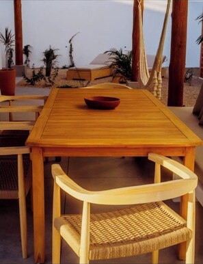 Een houten tafel met een schaal, omringd door rieten stoelen, buiten geplaatst met uitzicht op het strand en palmbomen.