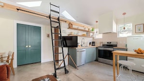 Una cocina compacta con televisión, estufa, horno y lavavajillas. Hay una escalera apoyada en la pared, una mesa de madera y una puerta verde.
