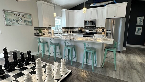 A modern kitchen with white cabinets, a central island with bar stools, a chessboard, and a sign on the wall. - Beachview Bliss at The Billfish Luxury 4BR Home in Gulfport (Gulfport)