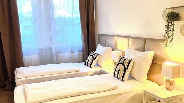 Une chambre d’hôtel avec deux lits, une grande fenêtre avec des rideaux, une table de chevet avec une lampe et une plante suspendue.