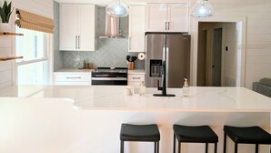 Een moderne keuken met witte kasten, een roestvrijstalen koelkast, een inbouwoven en een marmeren aanrecht. Er staan drie zwarte barkrukken bij het eiland.
