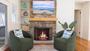 Een woonkamer met een stenen openhaard, twee groene fauteuils, een vloerkleed en een schilderij boven de openhaard.