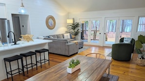 Een ruime woonkamer met een houten eettafel, een grijze bank en een keuken met een koelkast en barstoelen.
