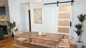 Een eetruimte met een houten tafel, een bank en stoelen. Er is een kroonluchter, een kaart aan de muur en een schuifdeur in boerderijstijl.