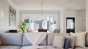 Una sala de estar con un sofá seccional gris grande, una manta blanca y un cojín azul. En el fondo, se observa una mesa de comedor con sillas, y una ventana grande con vista a un edificio.