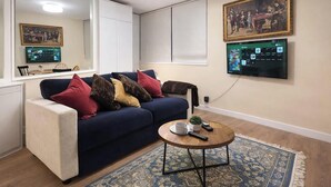 A modern living room with a dark blue sofa, multiple cushions, a round coffee table with a remote control, a small plant, and a decorative rug. - London Luxe Nest Your Perfect Short Stay (London)