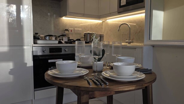 A modern kitchen with white appliances, a wooden table set with white dishes, and a glass vase. - London Luxe Nest Your Perfect Short Stay (London)