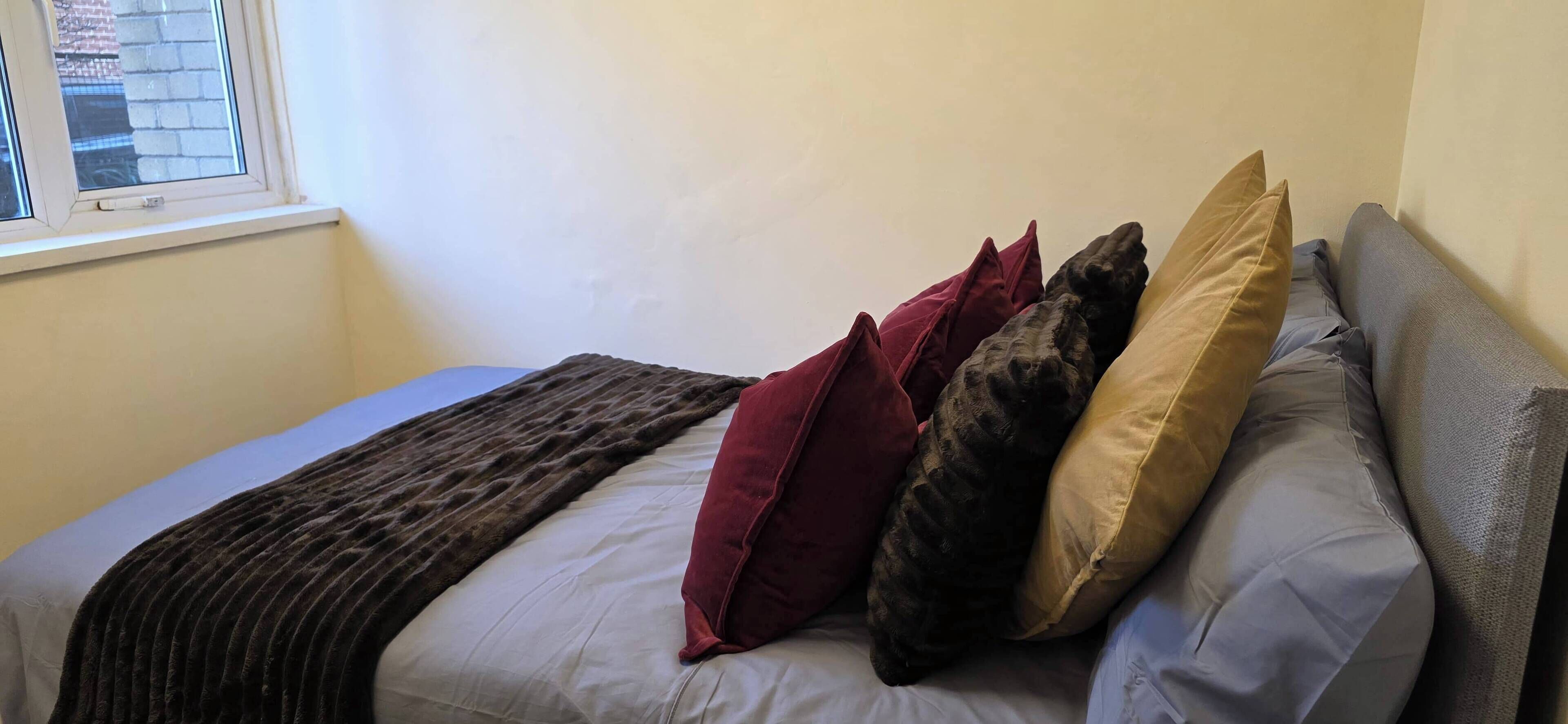 A bed with a dark brown blanket, multiple pillows in various colors, and a window with a view of buildings.