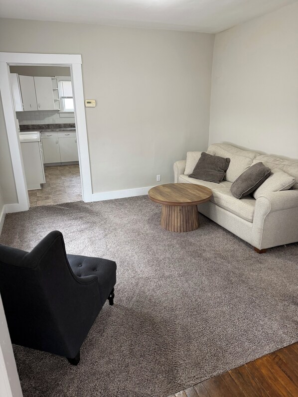 A living room with a beige sofa, a round wooden coffee table, and a black armchair. A kitchen is visible in the background.