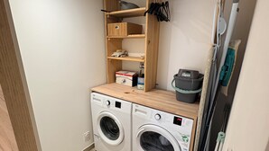Ein kleiner Waschbereich mit zwei Waschmaschinen, die nach vorne laden, einem Holzbodenregal darüber und einem Wäscheständer zur Rechten.