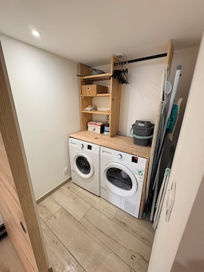 A compact laundry area with two front-loading washing machines, a wooden shelf unit above, and a drying rack to the right. - Village house near CNPE | Villebois (Villebois)