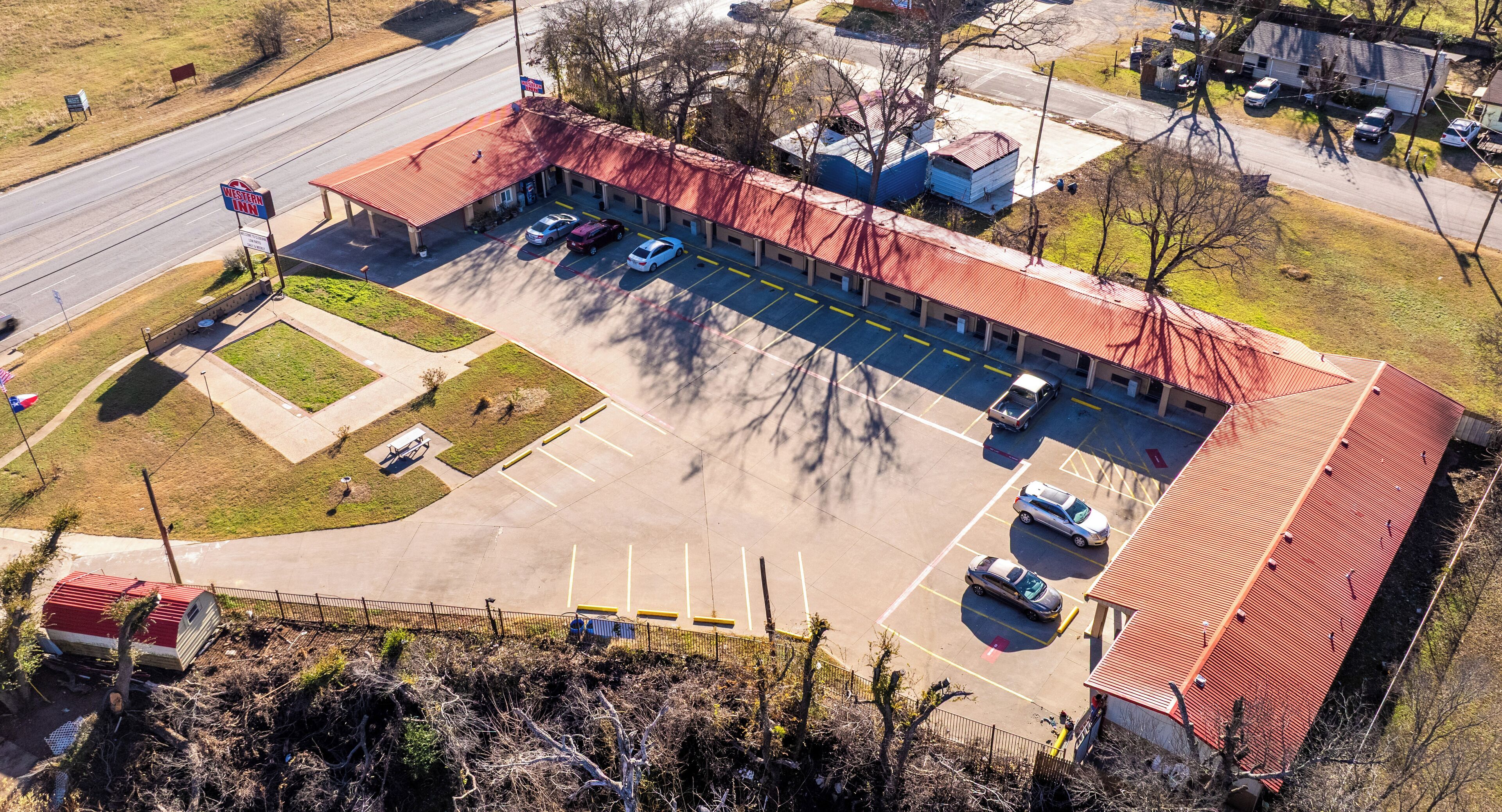 Vista aérea de un motel con techo rojo, múltiples espacios de estacionamiento y una pequeña área ajardinada con asta de banderas.
