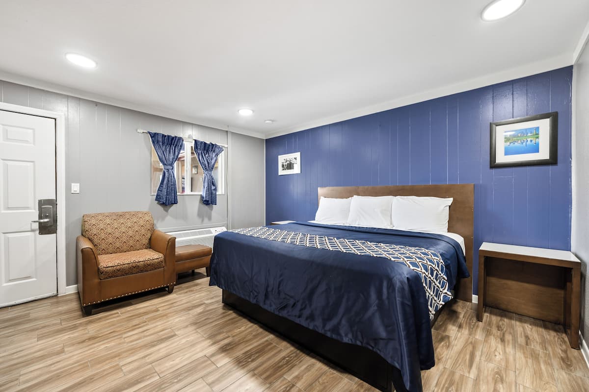 A bedroom with a bed, a chair, a nightstand, a window with curtains, and a framed picture on the wall.