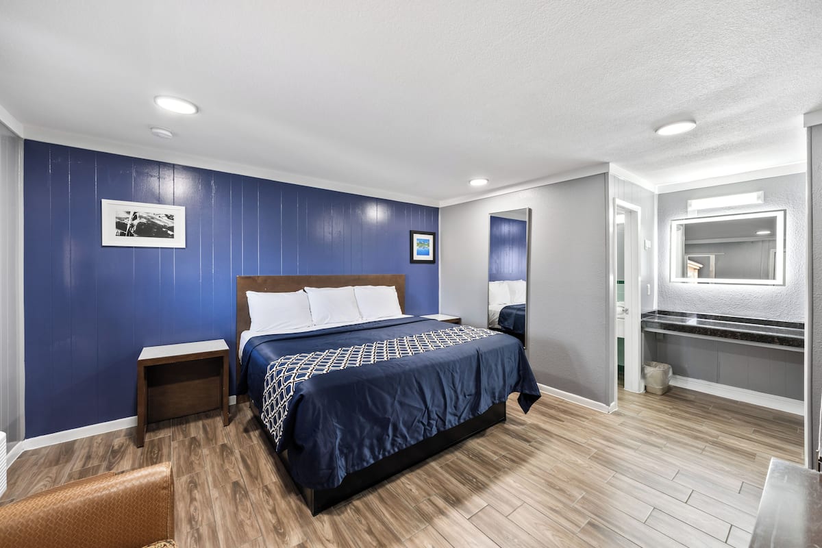 A modern hotel room with a large bed, wooden flooring, a small nightstand, a chair, and a bathroom with a mirror and sink.