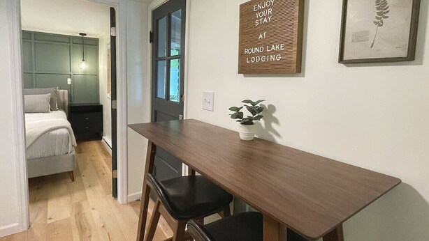 Une pièce de taille modeste, avec une table à manger en bois, deux chaises noires, une petite plante en pot, un lit avec une tête de lit grise et une porte donnant sur une autre pièce.