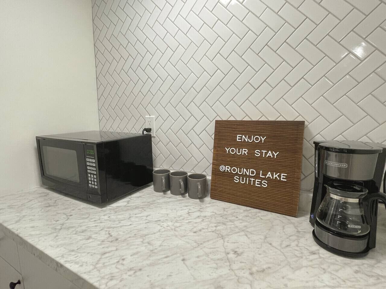 Una encimera de cocina con microondas, cuatro tazas, una cafetera y un cartel que dice "ENJOY YOUR STAY @ROUND LAKE SUITES".