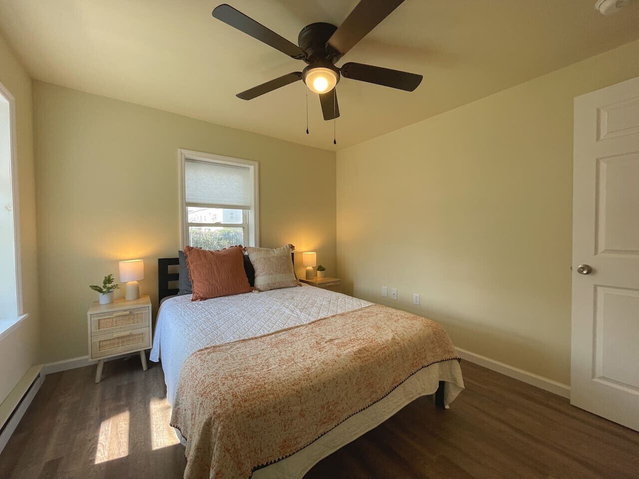 Une chambre à coucher comprenant un lit, des tables de chevet, un ventilateur de plafond, une fenêtre et une porte.
