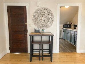 A wooden door, a wall-mounted decorative piece, a small table with two stools, and a glimpse of a kitchen area.
