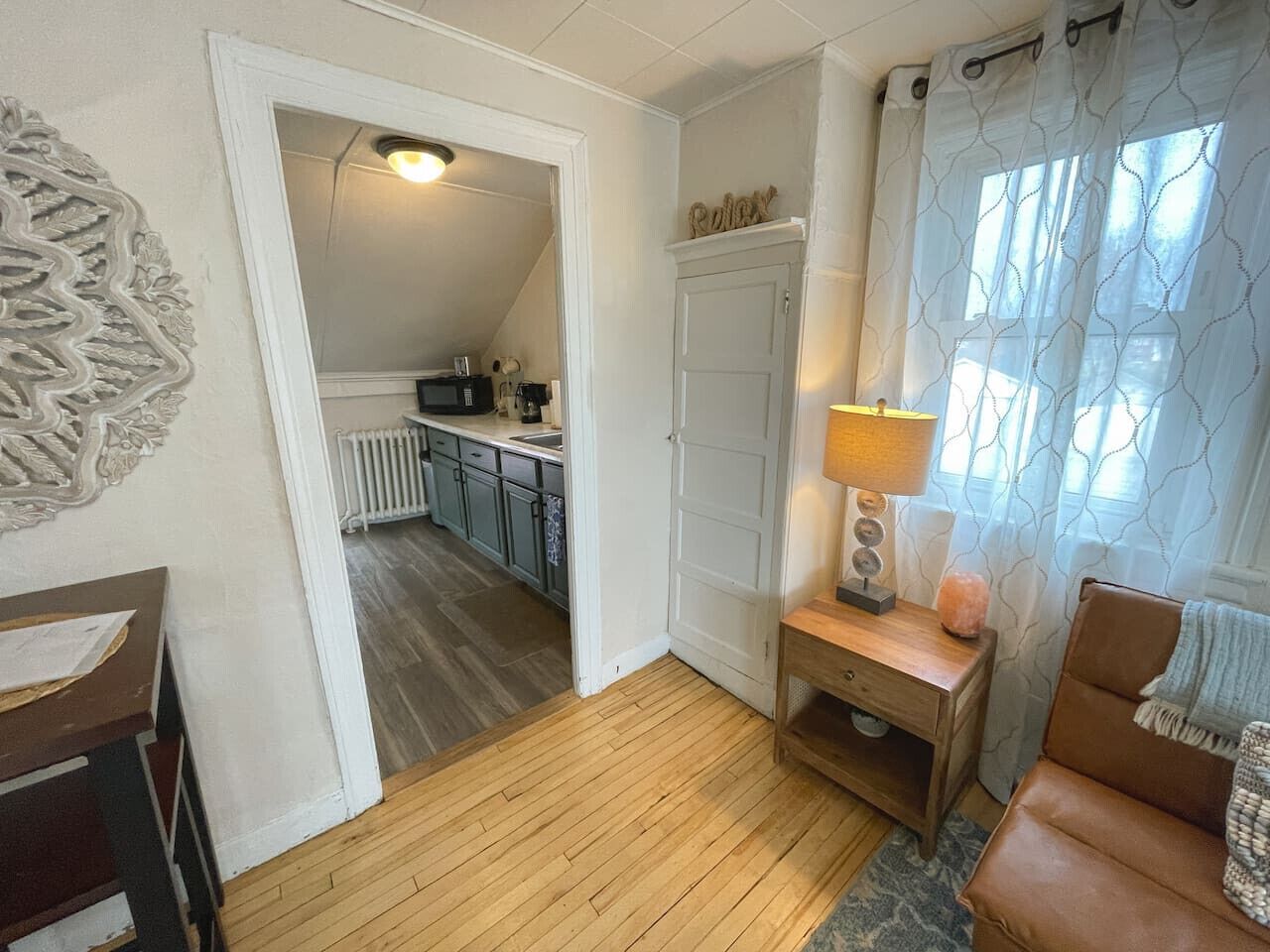 Un salon chaleureux avec un plancher en bois, un fauteuil en cuir marron, une petite table d’appoint en bois sur laquelle se trouve une lampe, et une vue sur la cuisine par une porte ouverte.