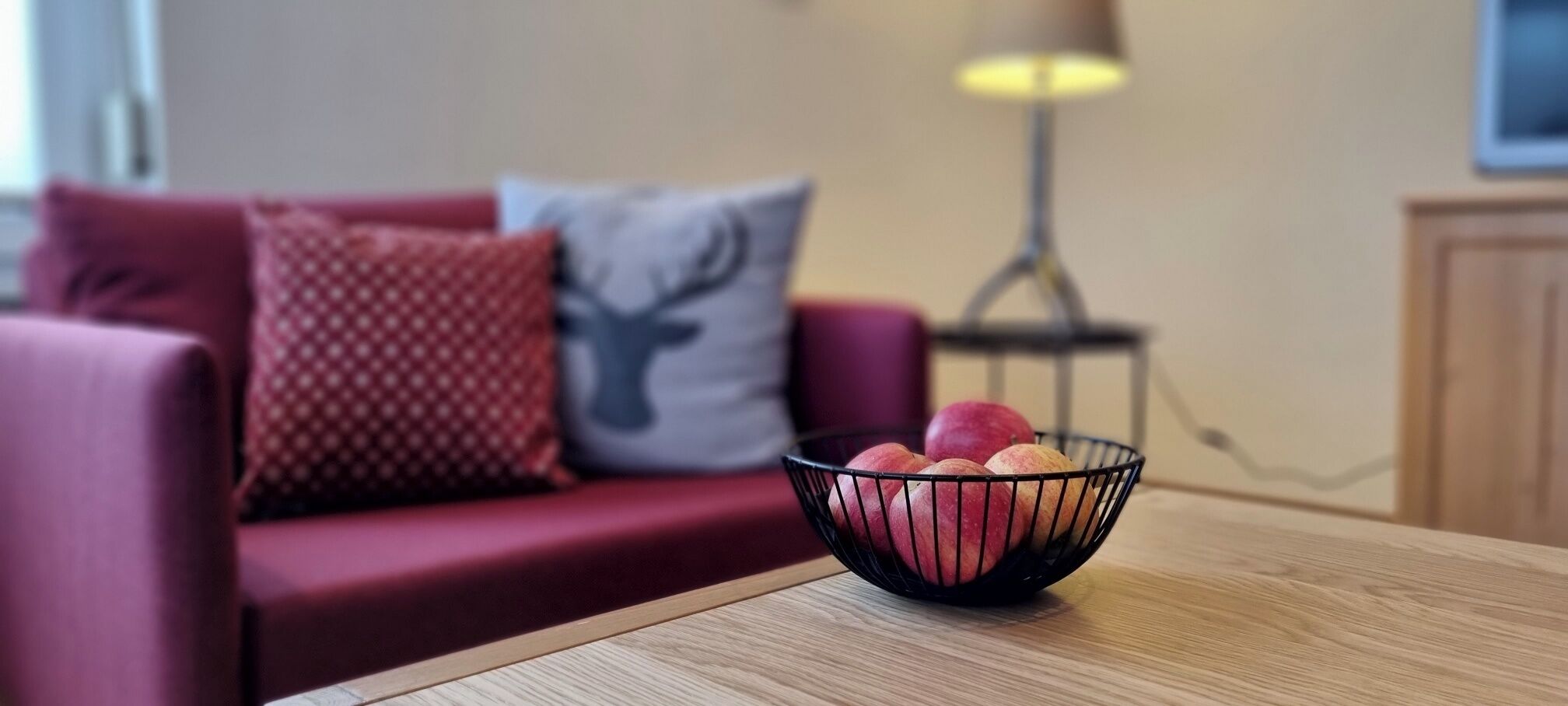A red sofa with patterned cushions, a black metal bowl with apples on a wooden table, and a lamp in the background.