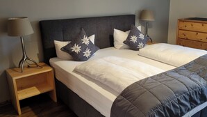 A neatly made bed with a grey and white duvet, two grey pillows with white floral patterns, a wooden nightstand with a lamp, and a wooden dresser with a black object on top. - Quiet vacation apartment with terrace and garden - wellness use included. (Reutte)
