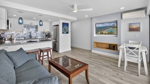 Ein modernes Wohnzimmer mit einer Couch, einem Couchtisch und Sesseln, eine Küche mit einem Edelstahl-Kühlschrank und ein großes Fenster mit Meerblick.