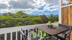 A balcony with wicker furniture and a potted plant, overlooking a lush green landscape and mountains. - Sacred Jungle Hideaway Surf Villa with AC (Princeville)