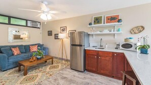 A compact living space with a kitchenette, a ceiling fan, a sofa with cushions, a coffee table, and a patterned rug. - Sacred Jungle Hideaway Surf Villa with AC (Princeville)