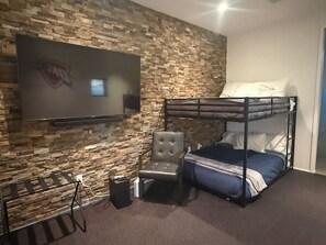 A room with a bunk bed, a flat-screen TV mounted on a stone wall, a cushioned chair, and a small speaker.