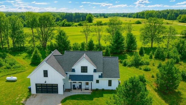 Una casa de dos pisos con un garaje, rodeada de campos verdes y árboles exuberantes.